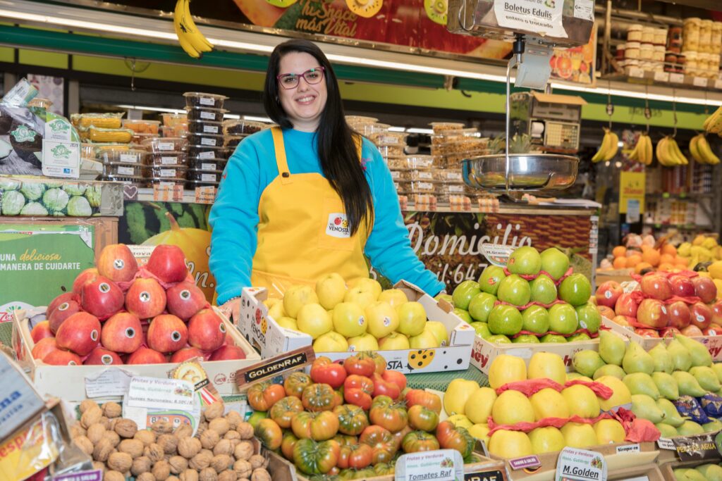 Smiling woman in a fruit market in Madrid, surrounded by fresh produce.